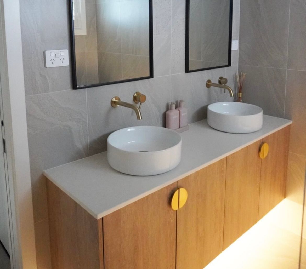 Bathroom with double vanity; wood cabinet, white countertop, round sinks, gold faucets and handles, and two black-framed mirrors. — Hallam & Sons in Beard, ACT
