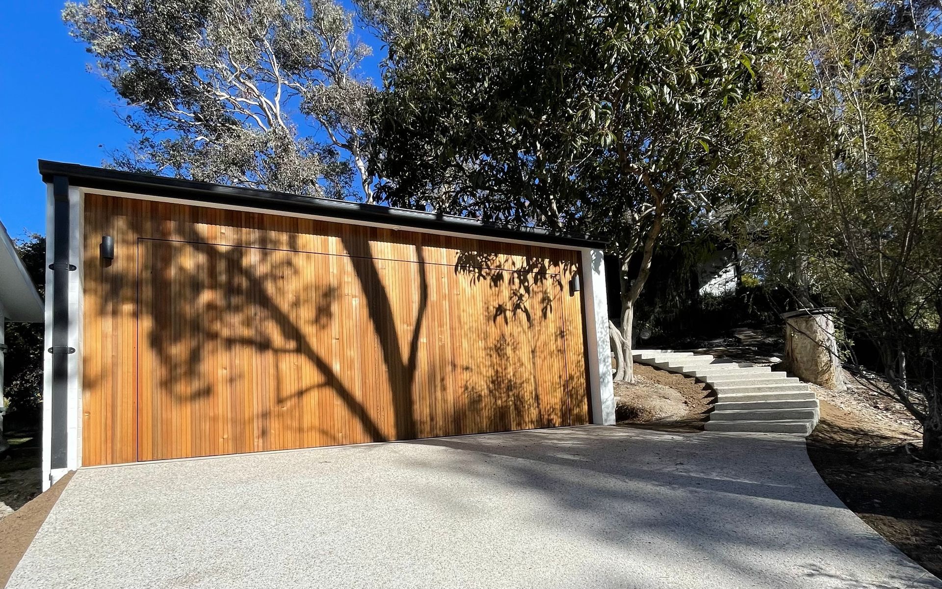 Wooden garage with gravel driveway, shadow of tree, path leading uphill. — Hallam & Sons in Beard, ACT