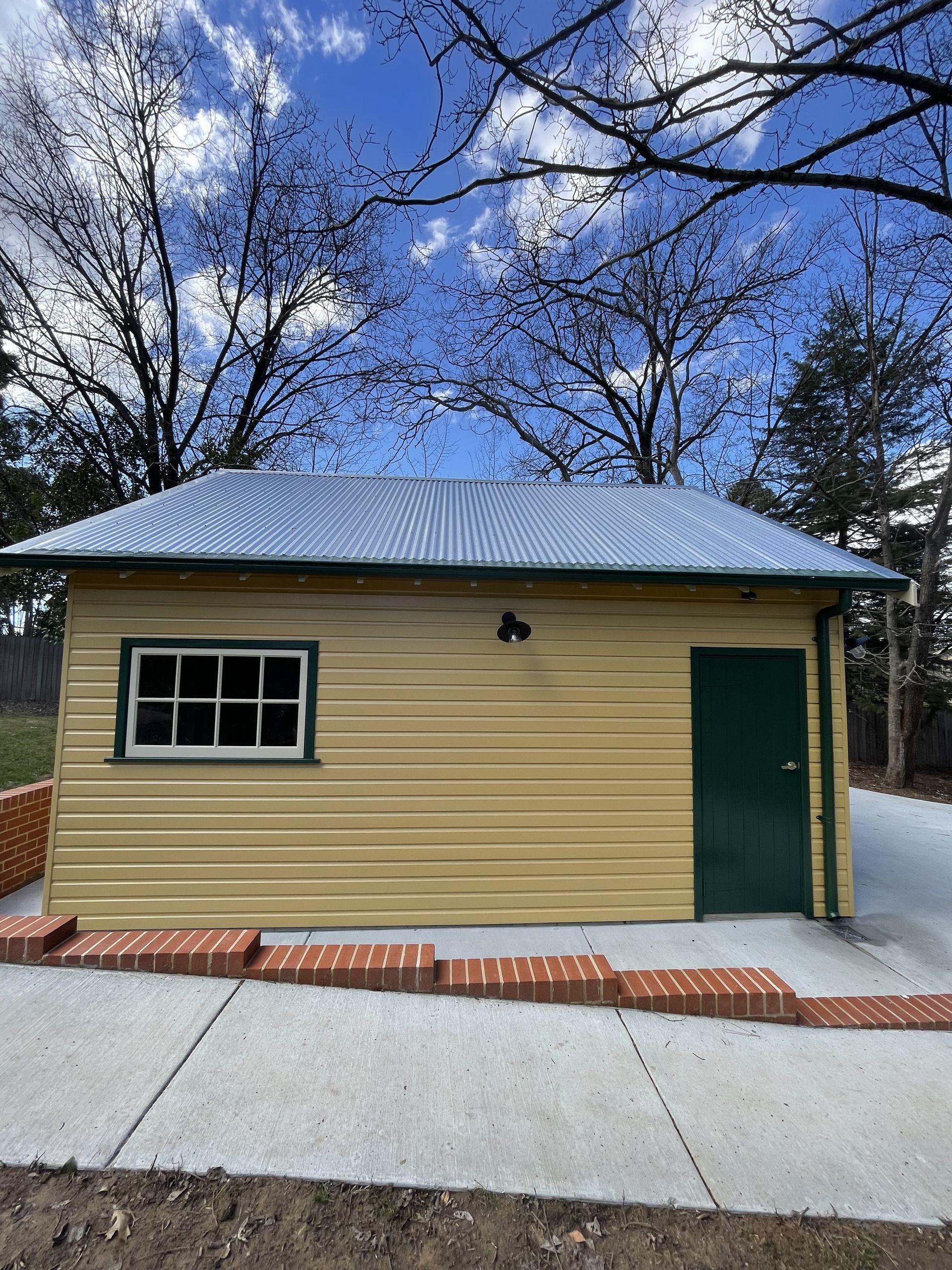 Yellow building with corrugated metal roof, green door and trim, small window, set against a blue sky with bare trees. — Hallam & Sons in Beard, ACT