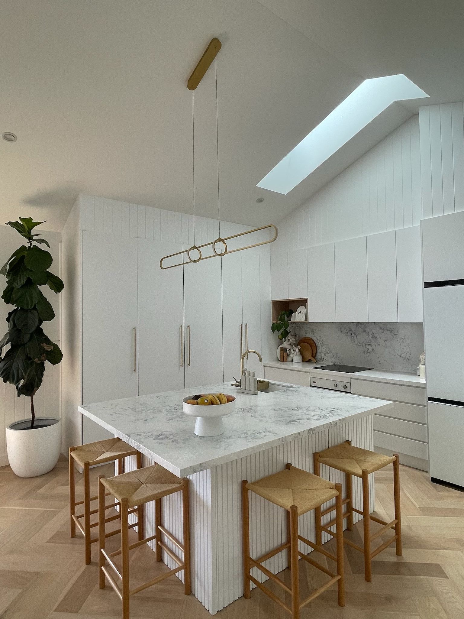 Modern white kitchen with island, pendant light, wood stools, skylight, and large plant. — Hallam & Sons in Beard, ACT