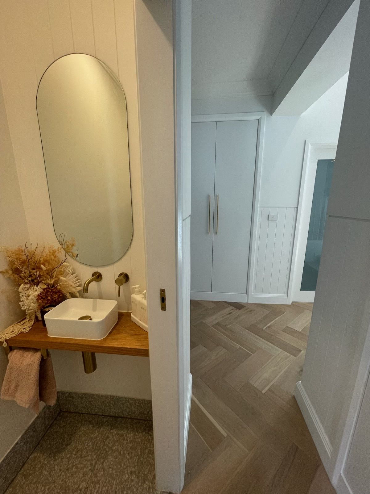 Bathroom with wooden counter, oval mirror, and herringbone floor. White walls, a closed door, and hallway visible. — Hallam & Sons in Beard, ACT