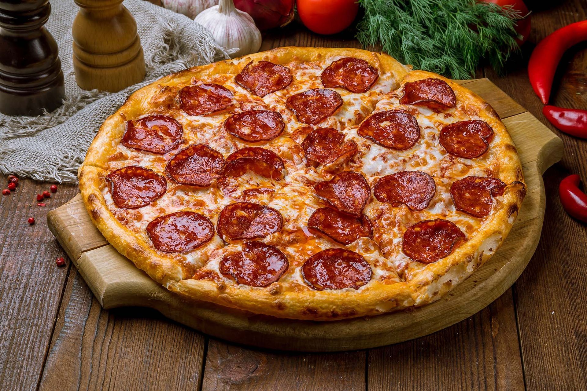 Pepperoni pizza on a wooden board with herbs and vegetables in the background