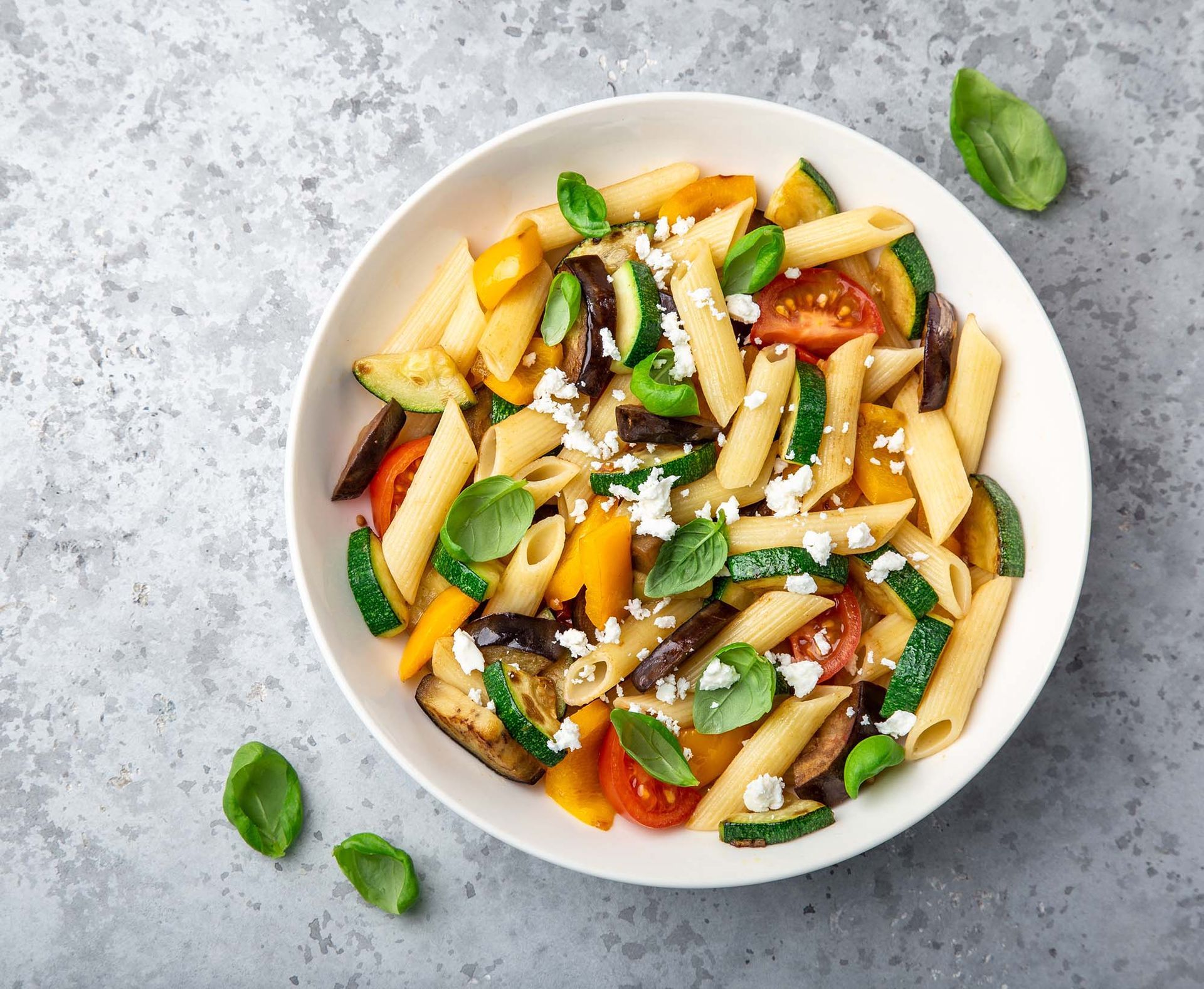 Bowl of penne pasta with tomatoes, basil, and cheese on a gray surface