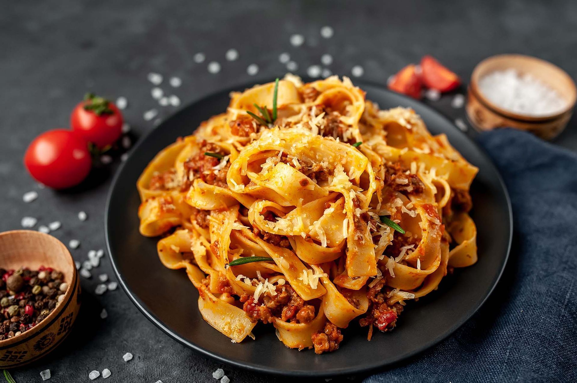 Pasta dish with tomato sauce and grated cheese on a black plate, surrounded by tomatoes and spices