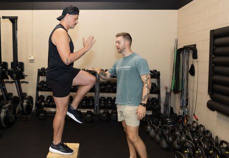Two men in a gym demonstrating a step-up exercise with a balance box and dumbbells nearby