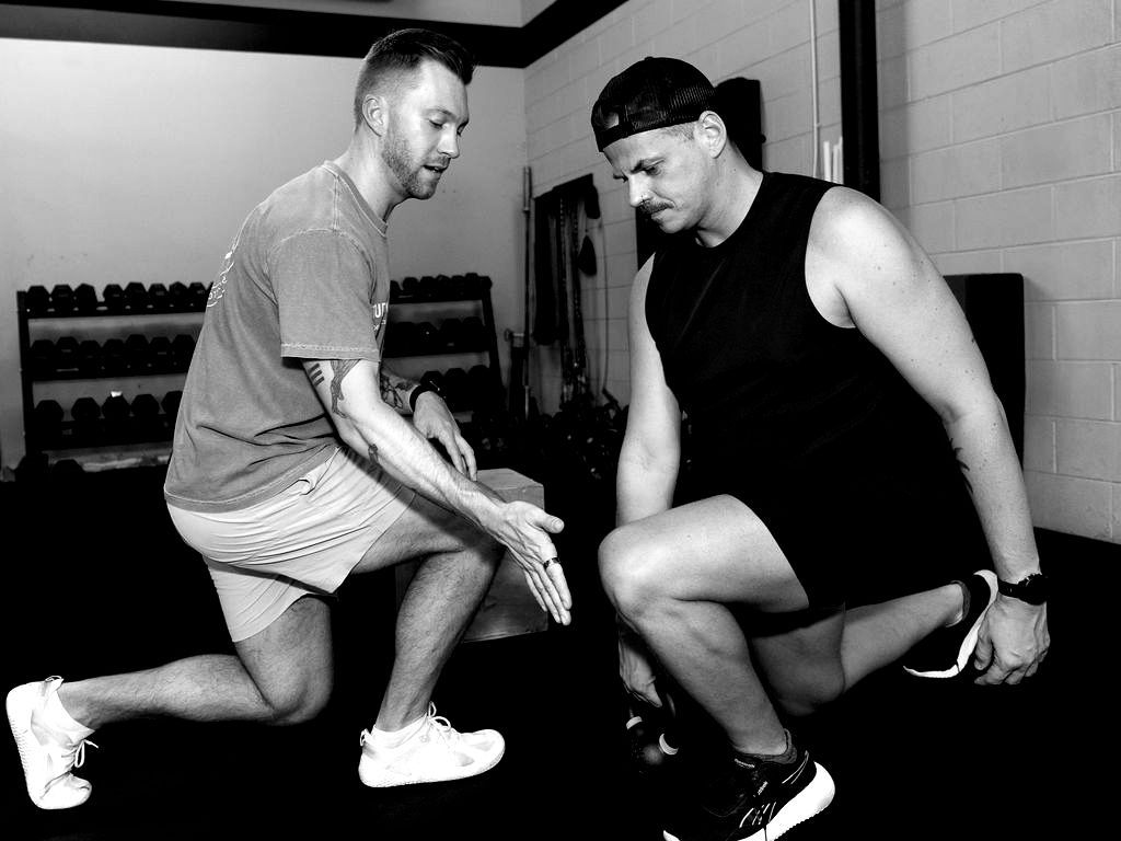 Two men doing a kneeling lunge stretch in a gym, one in a gray shirt and one in black.
