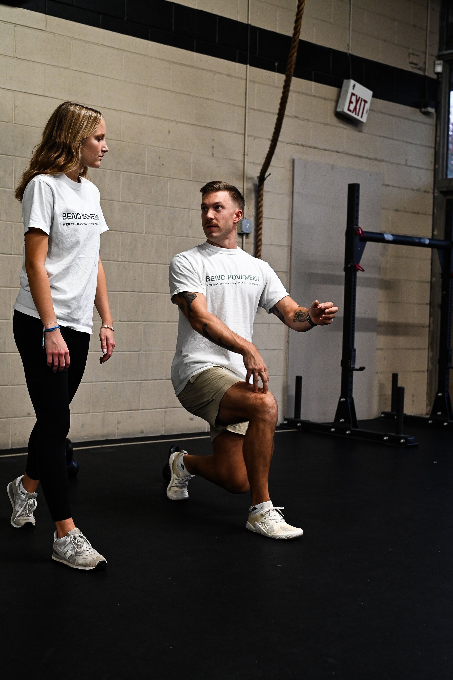 Trainer guiding a squat lunge in a gym, with a woman observing nearby.