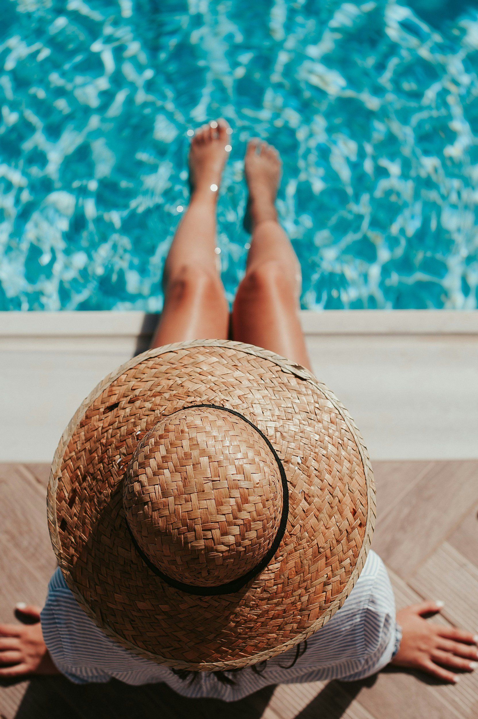 A woman wearing a straw hat is sitting on the edge of a swimming pool.