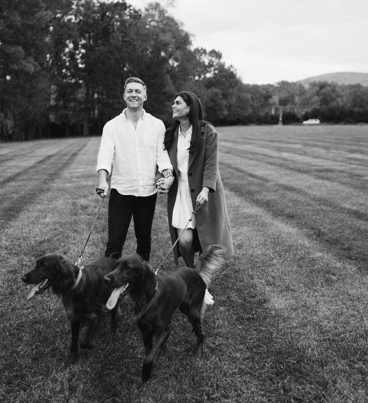 Two people walking three dogs on a grassy field, with trees in the background.