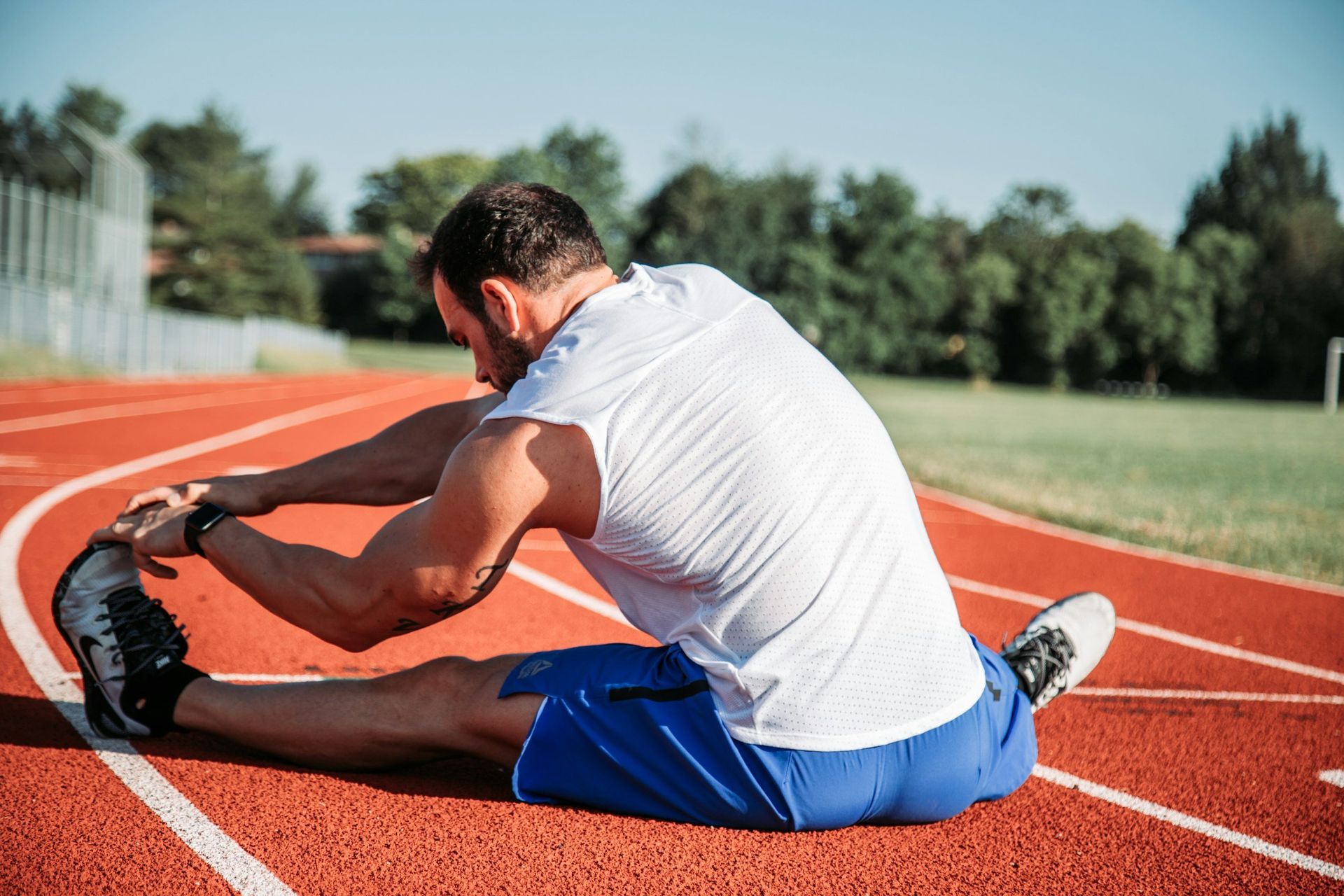 Runner stretching on a red track in blue shorts and a white shirt under the sun