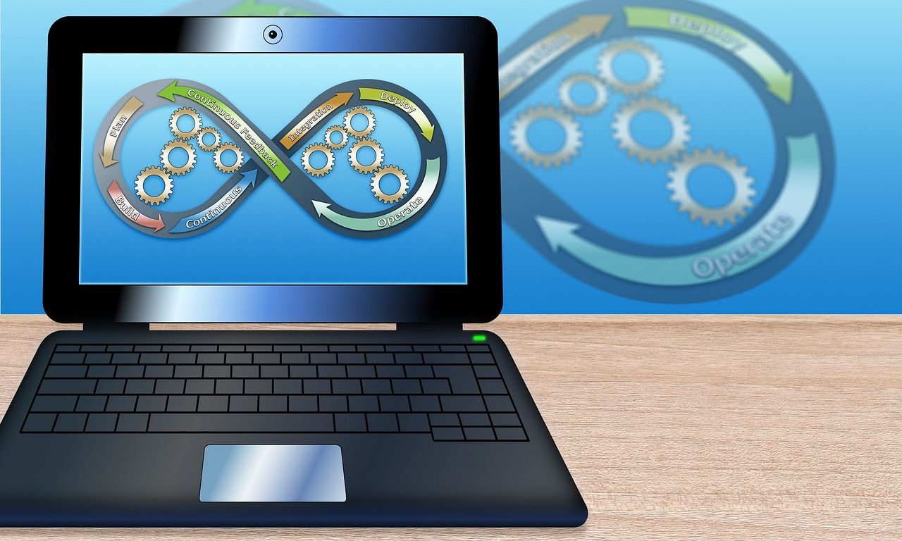 A laptop sits on a table, displaying an infinity loop diagram with interconnected gears representing a DevOps cycle.