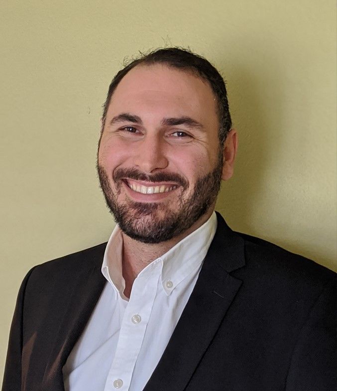 Man with dark hair and beard, smiling, wearing a white button-down shirt and black blazer, against a light yellow background.