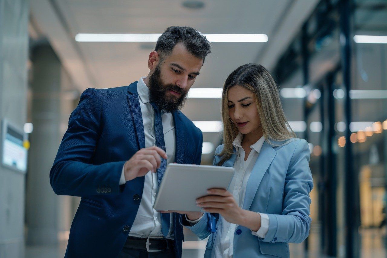 Man and woman in suits looking at a tablet in a hallway, discussing something.