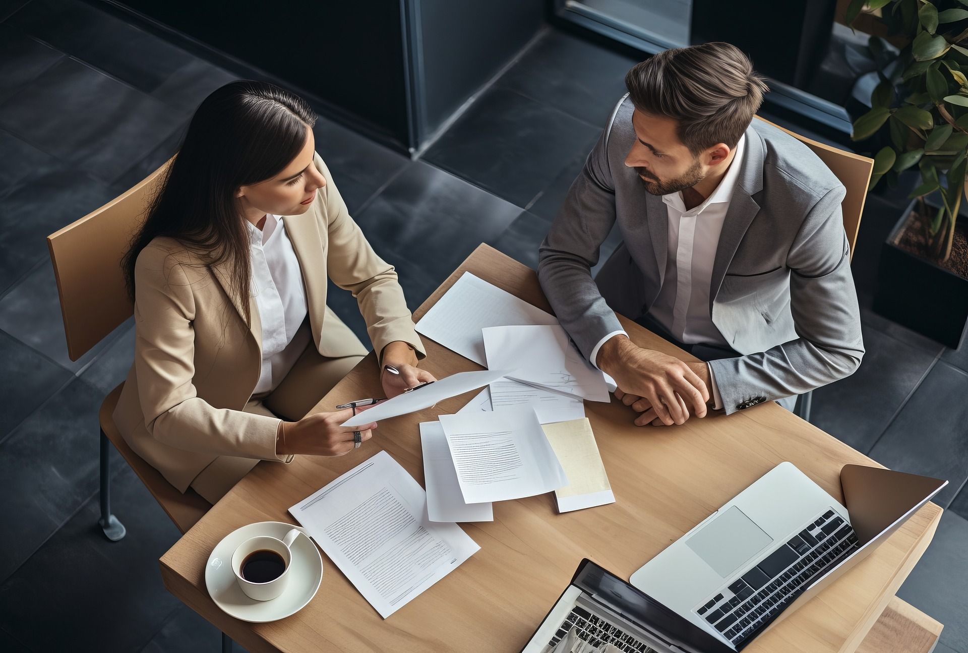 Man and woman in suits review documents at a table with a laptop and coffee.