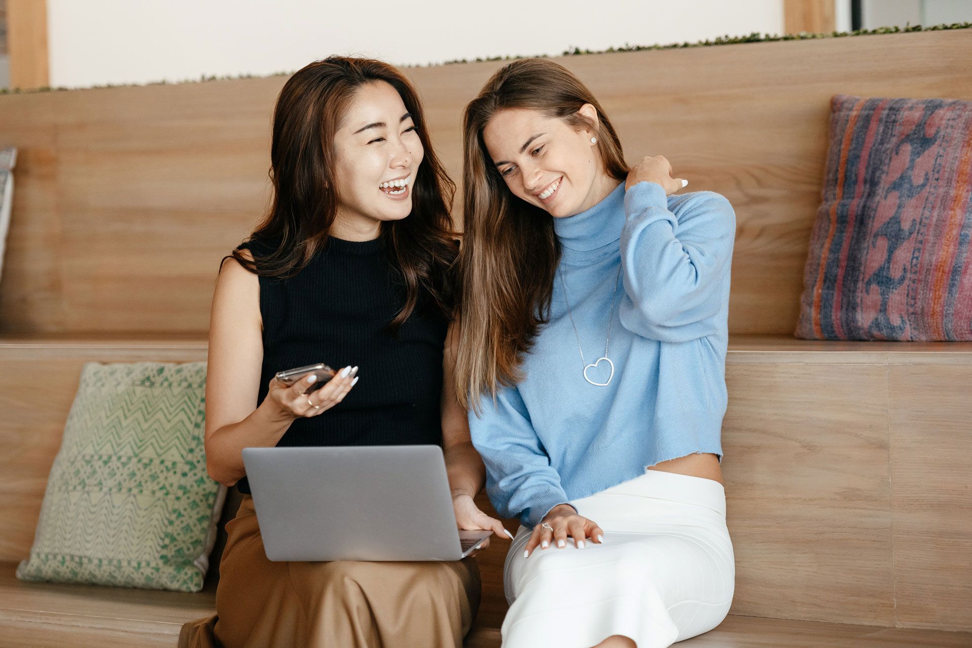 Two women sit on wooden bench, looking at a laptop. One holds a phone, the other touches her neck and smiles.