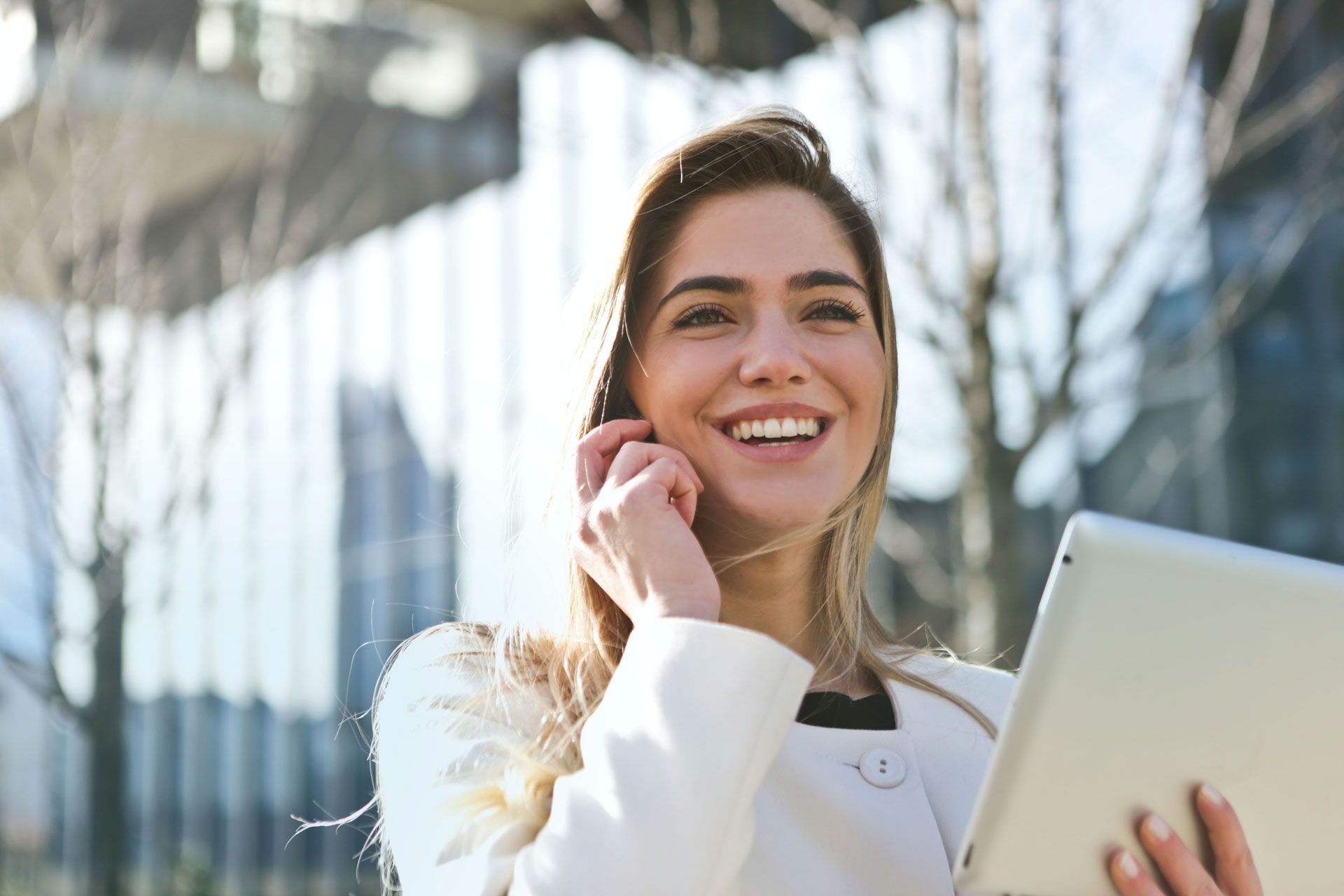 Woman smiles while holding a tablet and adjusting her earbud outside a modern building.