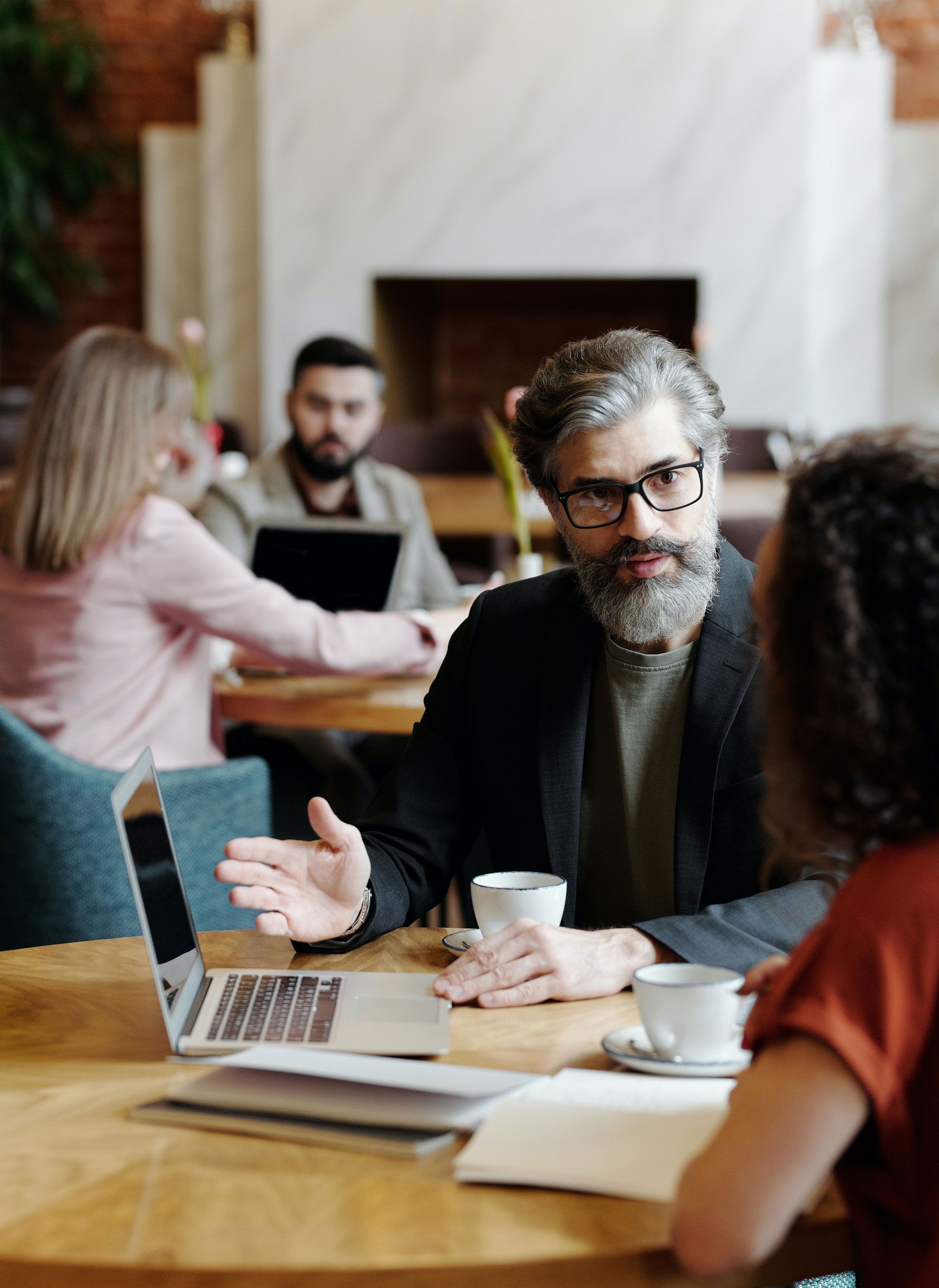 Man in glasses gestures at laptop while speaking to a person at a cafe table; others work nearby.