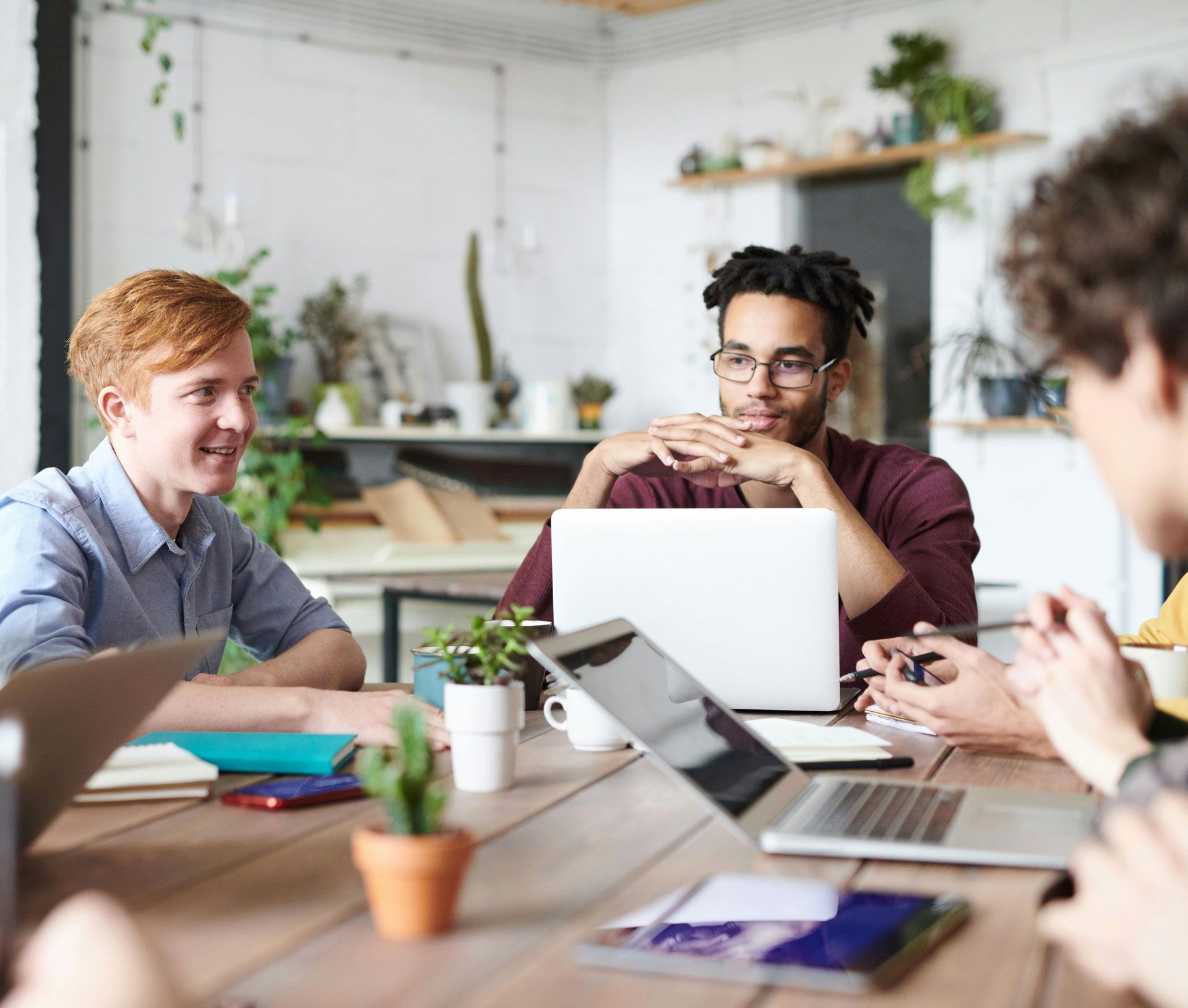 Four people at a wooden table, laptops open, having a discussion in a bright office setting.