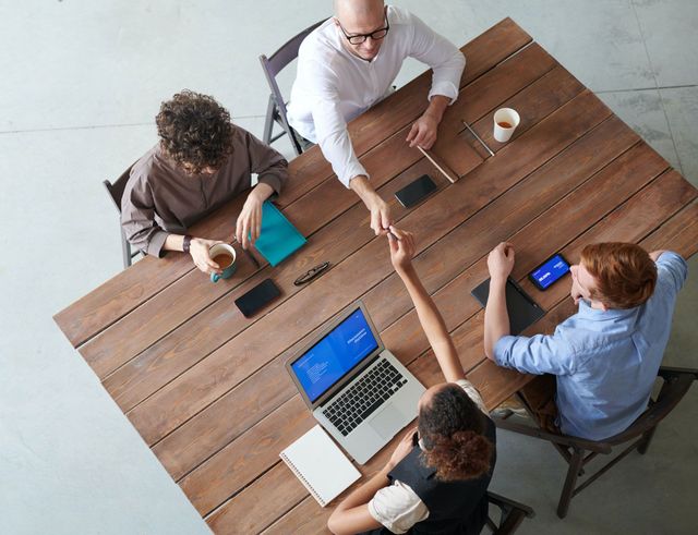 People in an office meeting around a wooden table, shaking hands. Laptop, notebooks, coffee cups are visible.