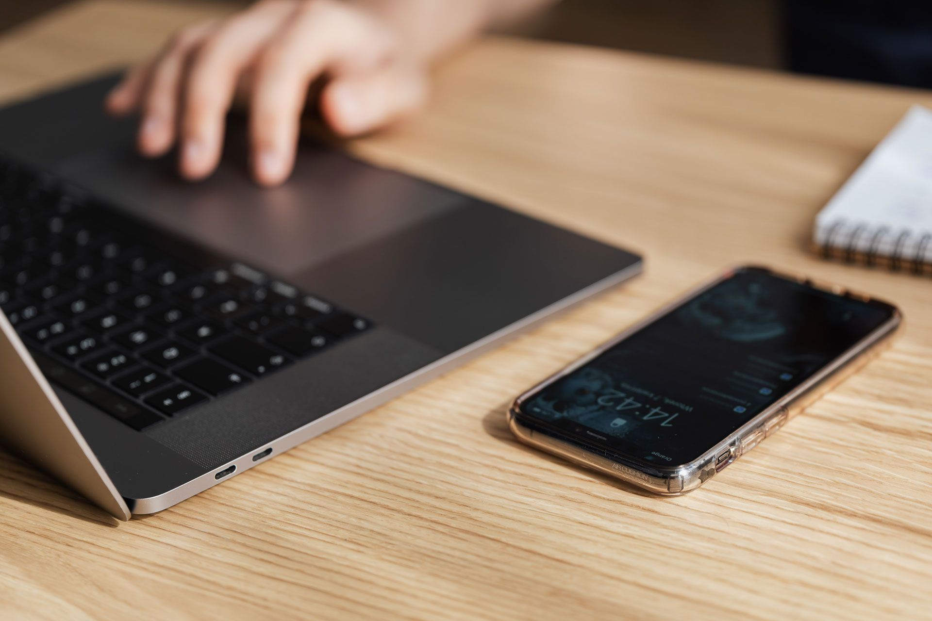 Laptop and phone on a wooden desk with a hand reaching towards the laptop.