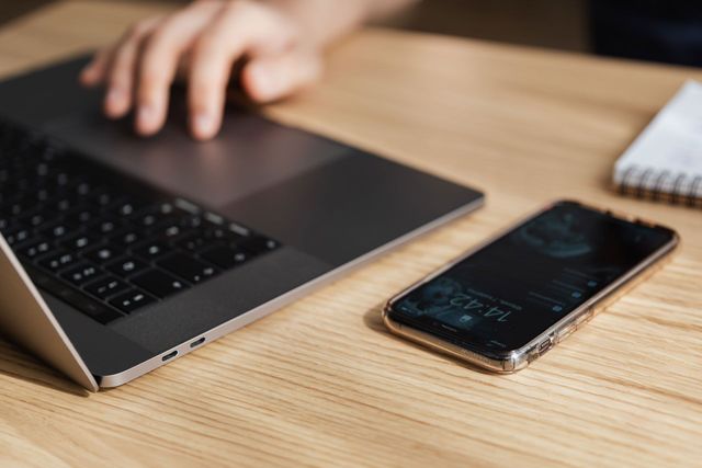 Laptop and phone on a wooden desk with a hand reaching towards the laptop.