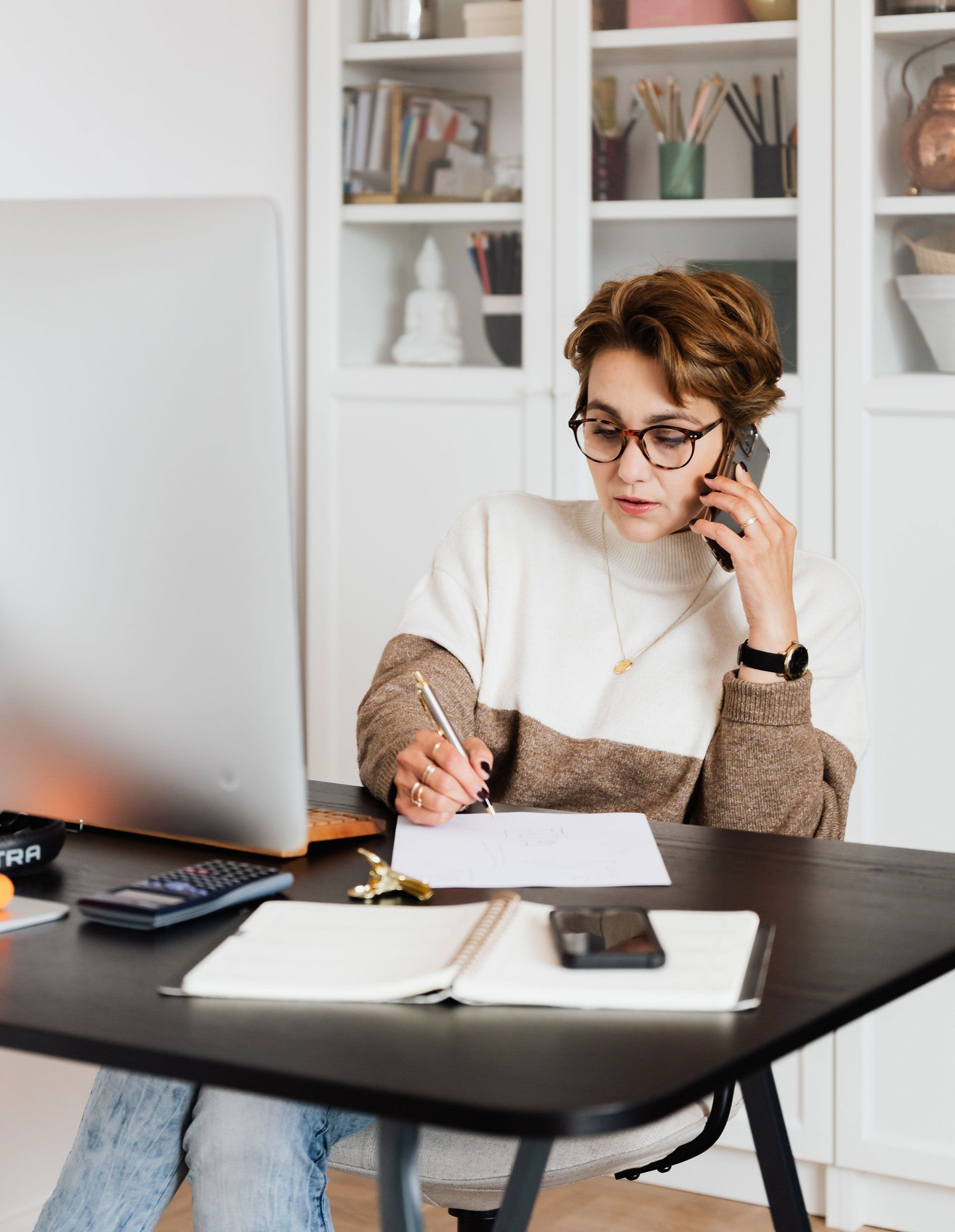 Woman on phone, writing at desk with computer, notebook, and phone. White sweater, glasses.