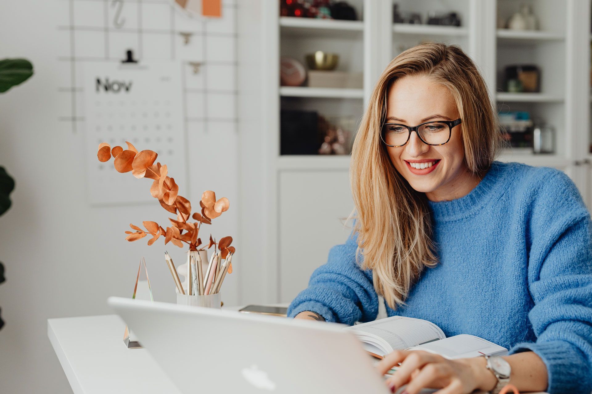 Woman with glasses smiles while working on a laptop at a white desk in a room.