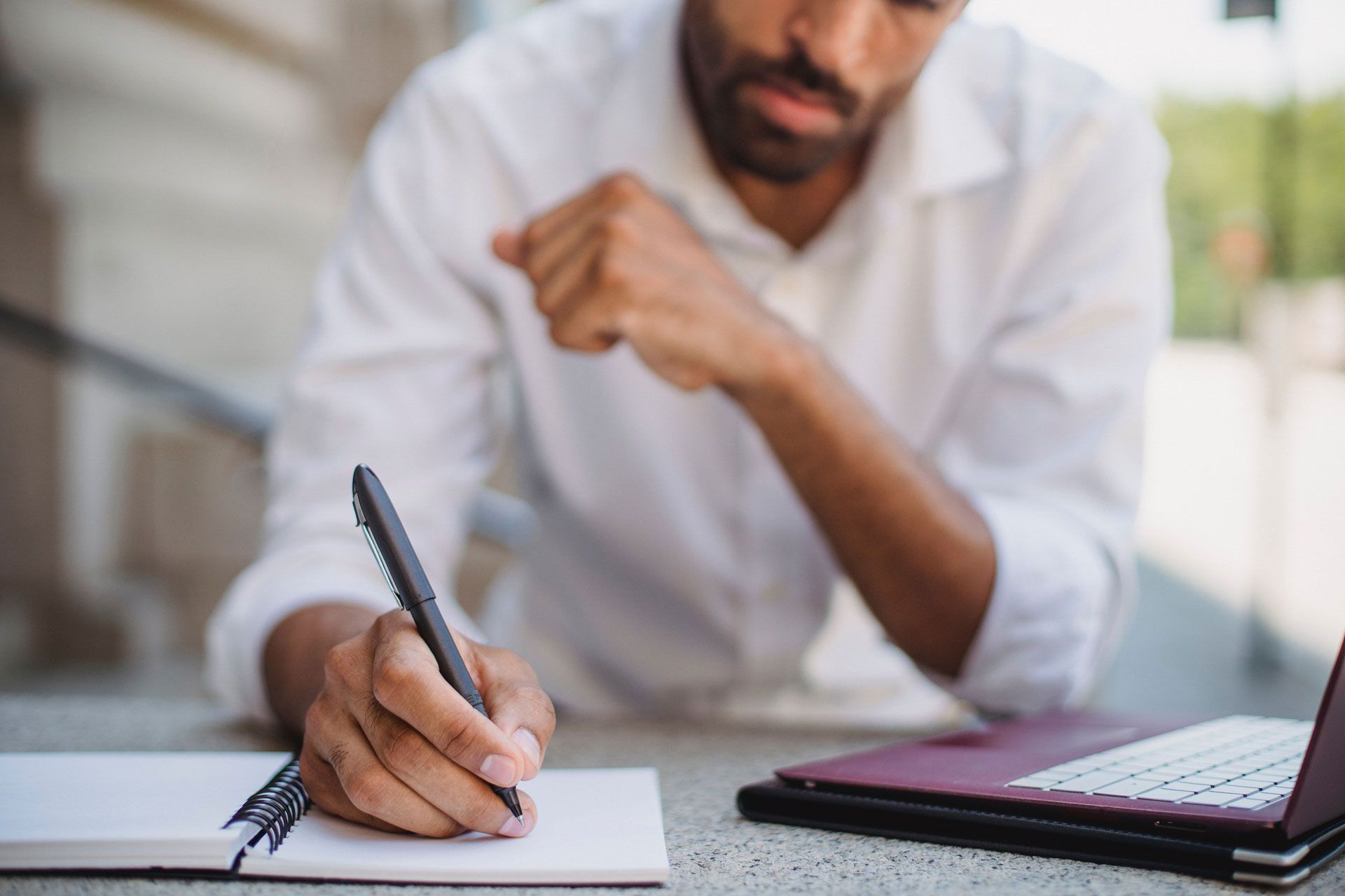 Man writing in notebook outdoors, laptop nearby, focused expression.