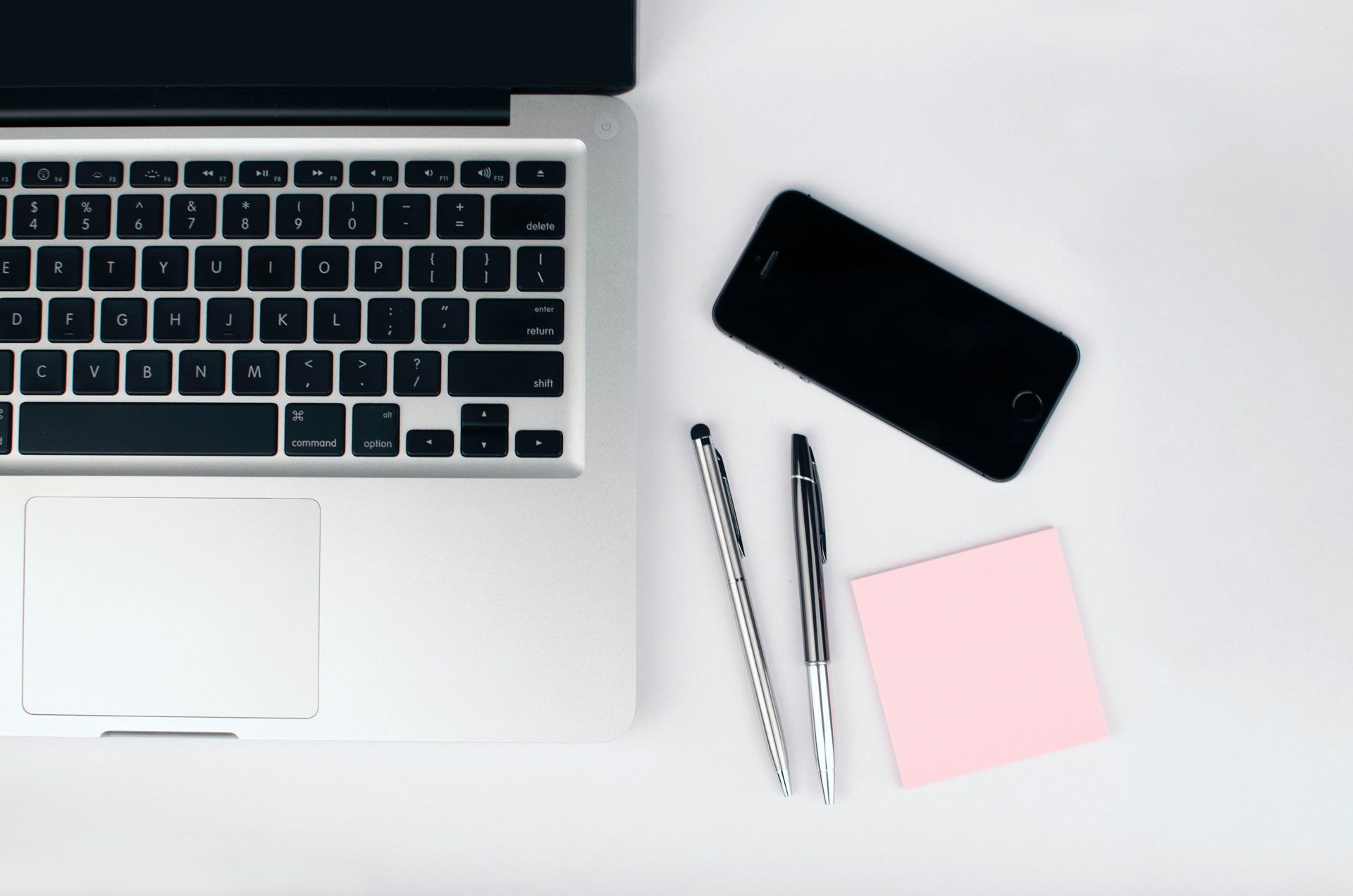 Laptop, smartphone, pens, and pink sticky note on a white surface.