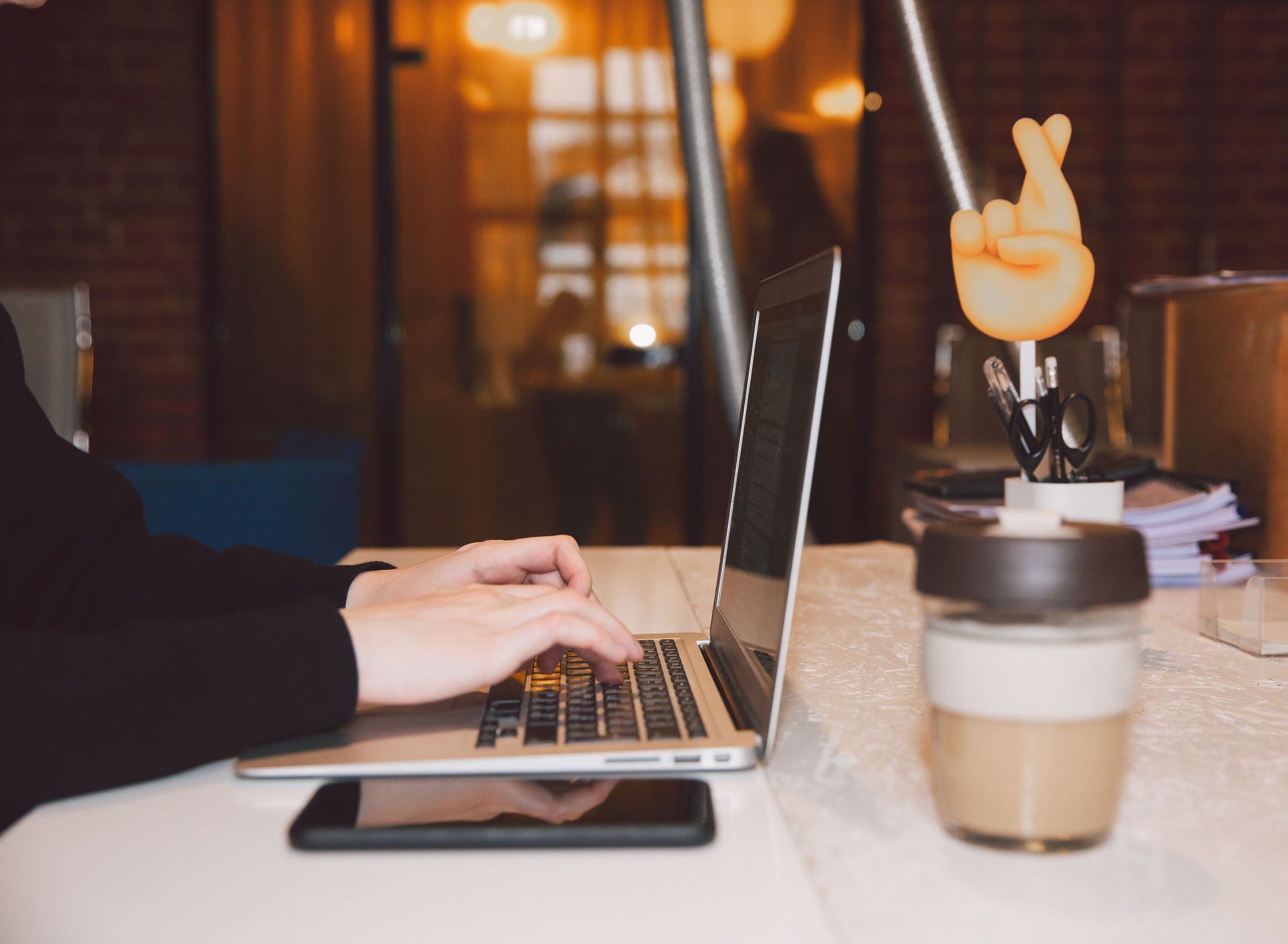 Person typing on a laptop at a desk with a coffee mug, phone, and decorative hand gesture.