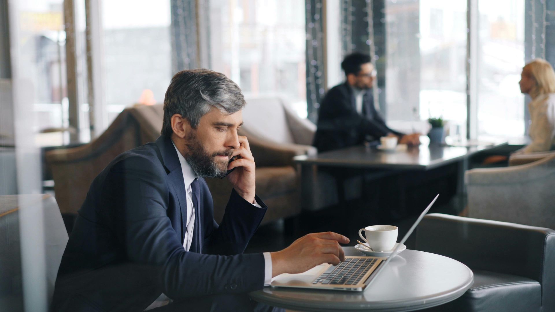 Man in a suit using a laptop and phone at a cafe table; other people seated in the background.