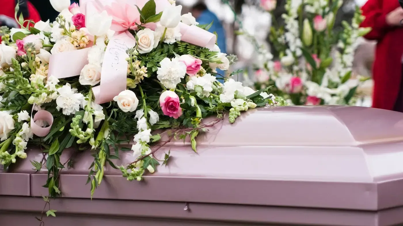 A pink casket covered in white and pink floral arrangements at an outdoor memorial service.
