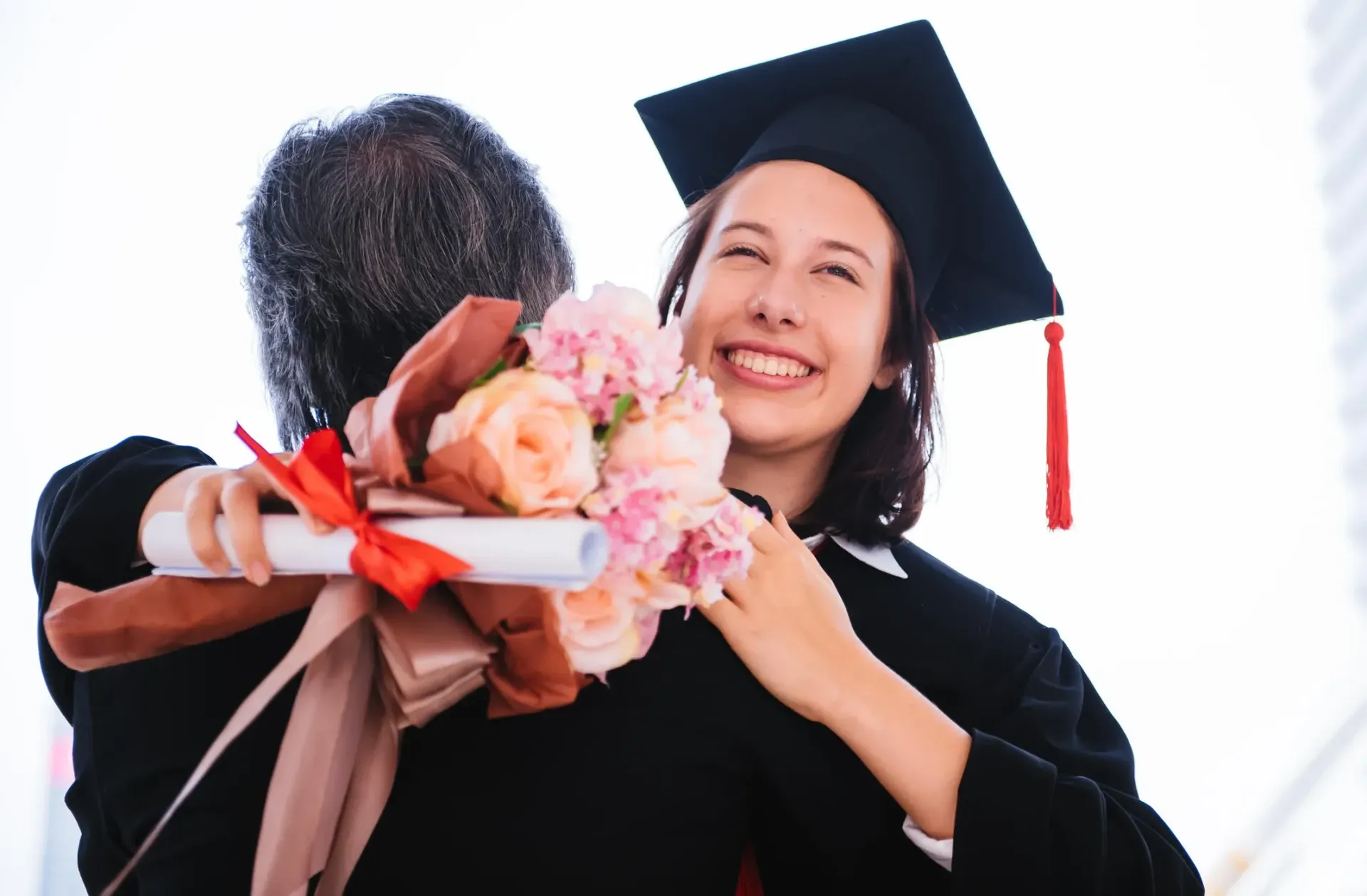 A woman in a graduation cap and gown is hugging a man holding a bouquet of flowers.
