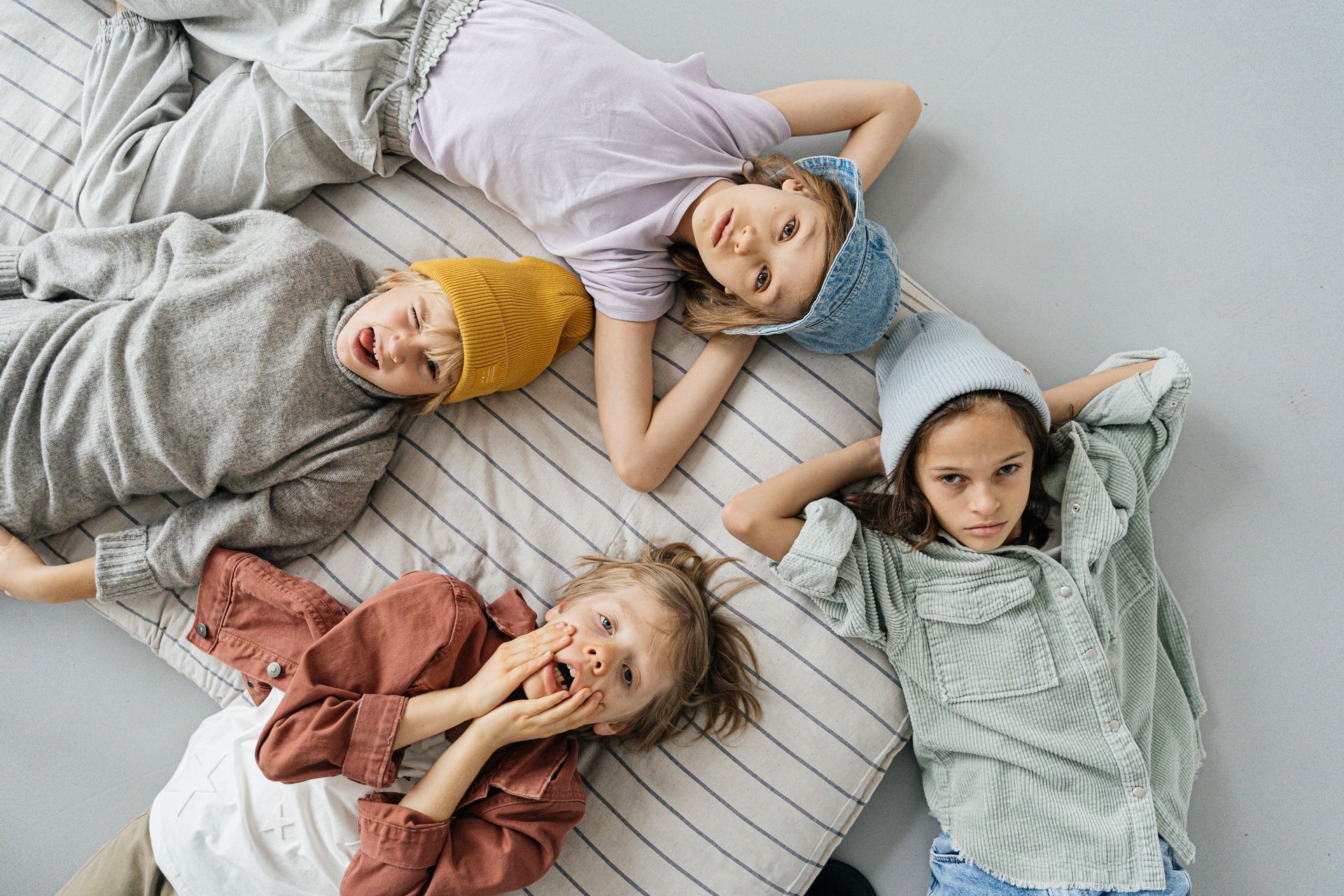 Vier kinderen liggen in kleurrijke kleren op een bed, kijken omhoog naar de camera en lachen.