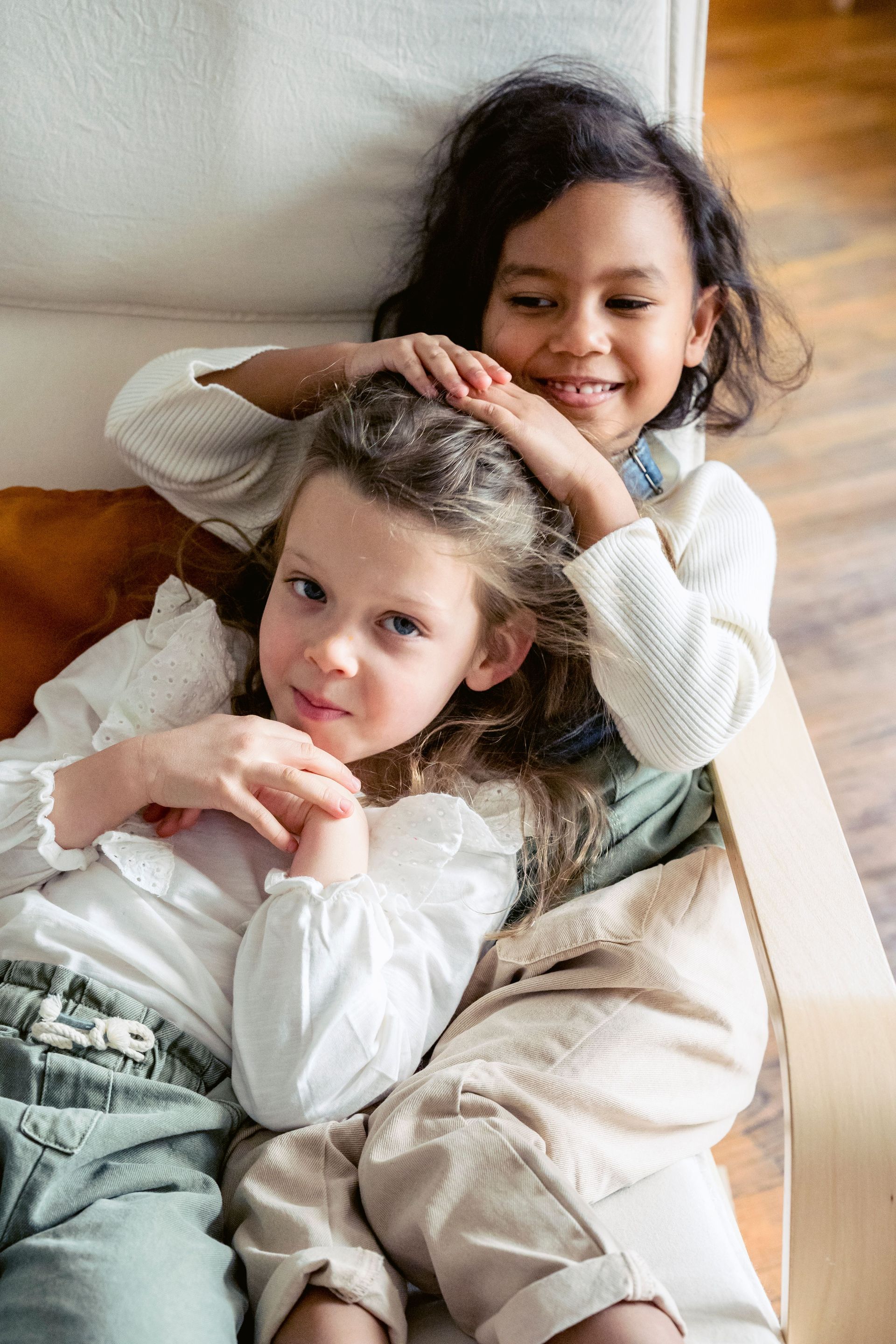 Twee kinderen liggen samen op een beige bank, lachend en knuffelend in het zachte, natuurlijke licht.