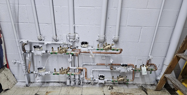 Valves and pipes mounted on a white cinder block wall, with concrete floor below.