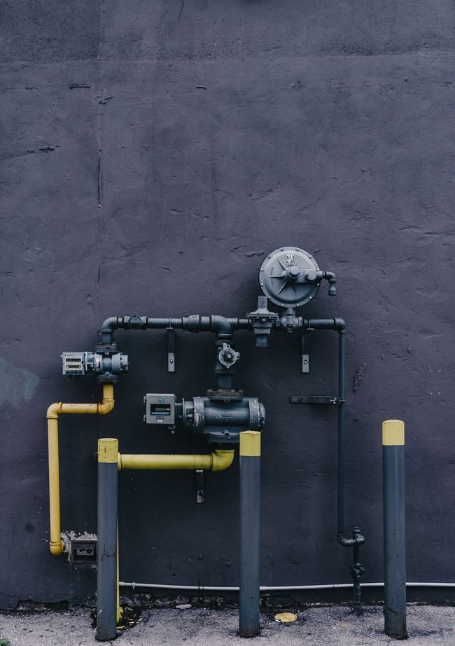 Industrial gas meter and pipes mounted on a dark wall, with yellow and black piping.
