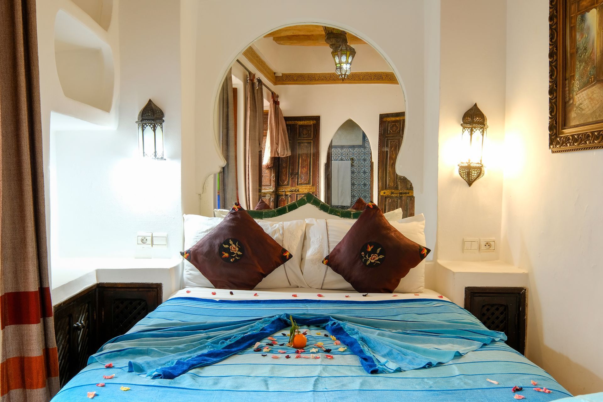 Bedroom with arched mirror, bed covered with blue fabric, decorative pillows, Moroccan-style lanterns.