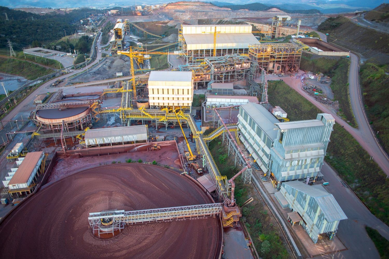 Vista aérea de um grande complexo industrial de mineração com instalações de processamento e lagoas de rejeitos, situado em uma paisagem montanhosa.
