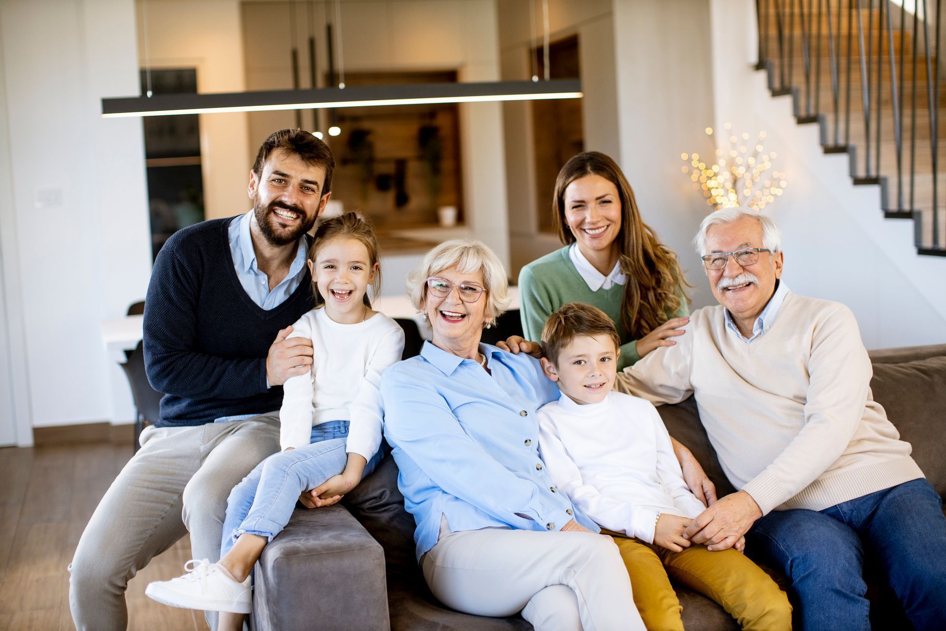 Família de sete pessoas sorrindo, sentadas juntas em um sofá na sala de estar.