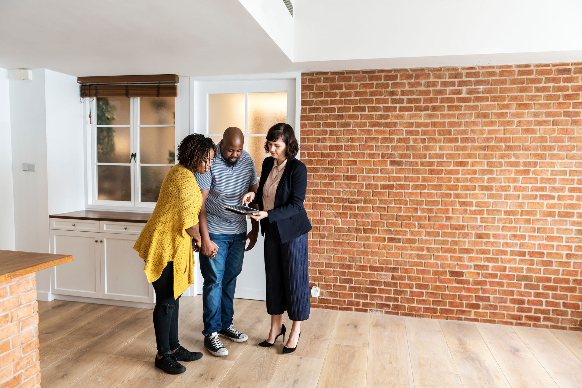 Real estate agent showing a couple a tablet in a new apartment with brick wall.