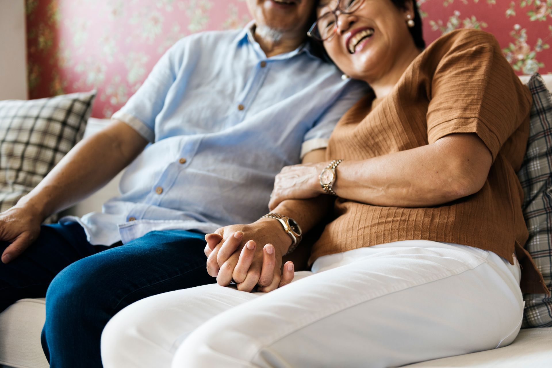 Elderly couple, smiling, holding hands on a couch, with woman leaning on the man's shoulder.