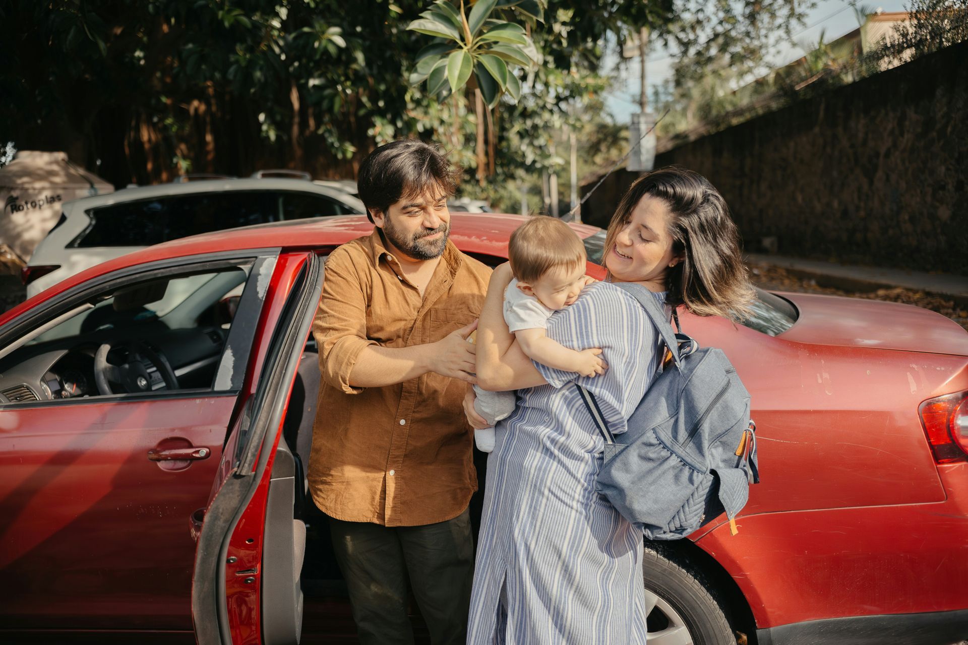 Family of three beside a red car, mother holding baby, father smiling, outdoor setting.