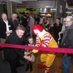 A mcdonald 's clown is cutting a pink ribbon in front of a crowd of people.