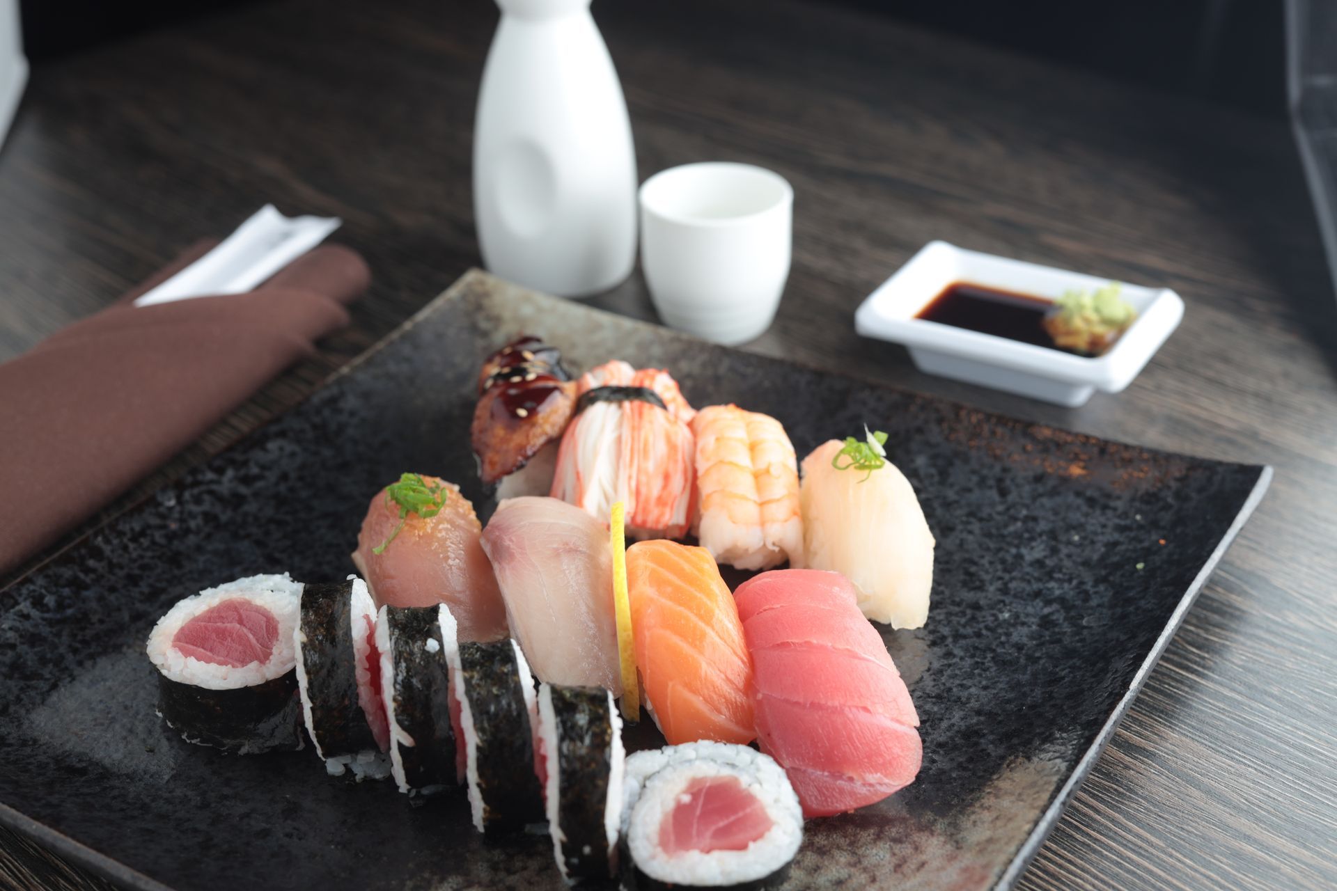 Sushi assortment on a black plate with chopsticks, soy sauce, and tea on a dining table
