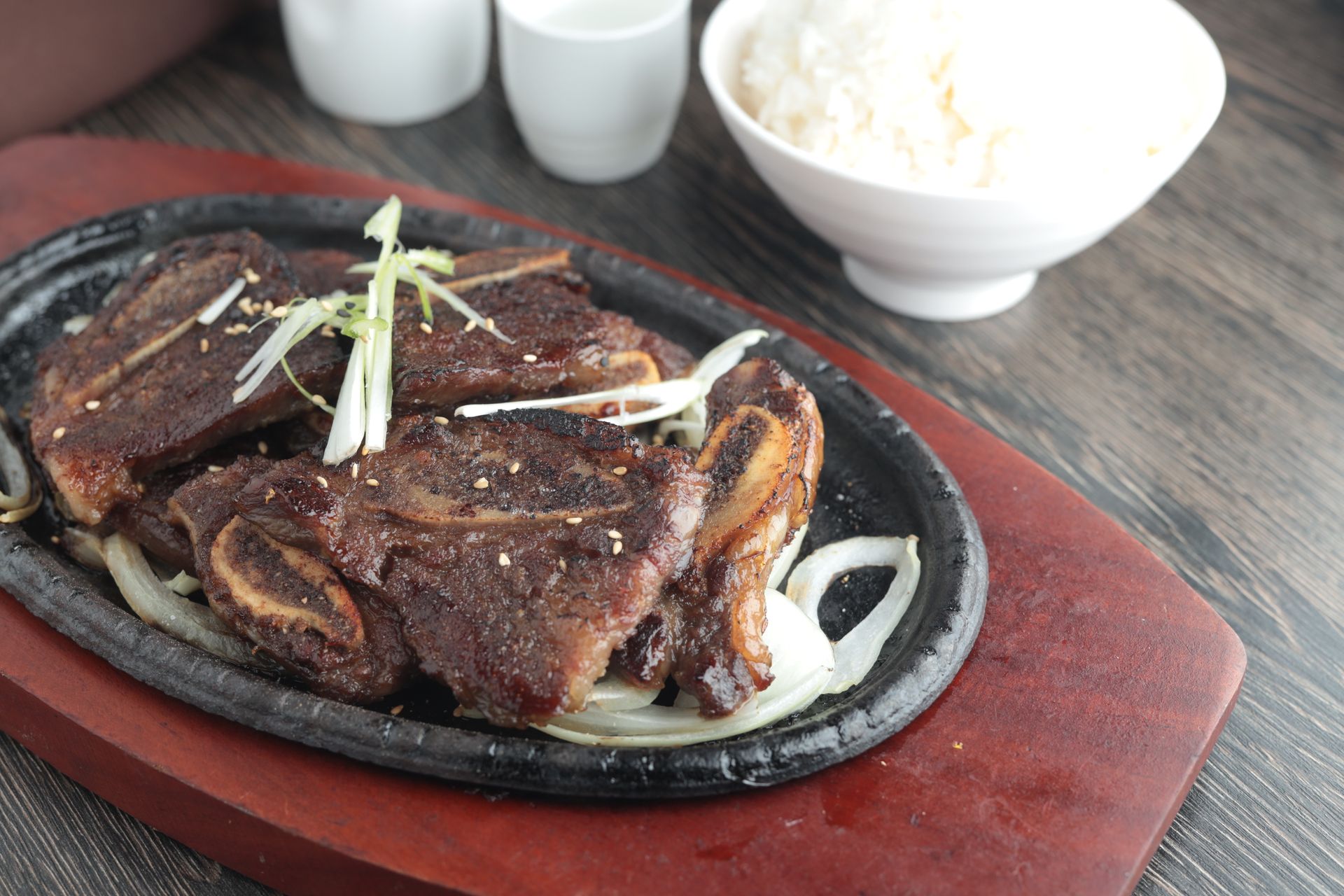 Grilled short rib on a sizzling plate with onion garnish and a bowl of rice on a wooden tray