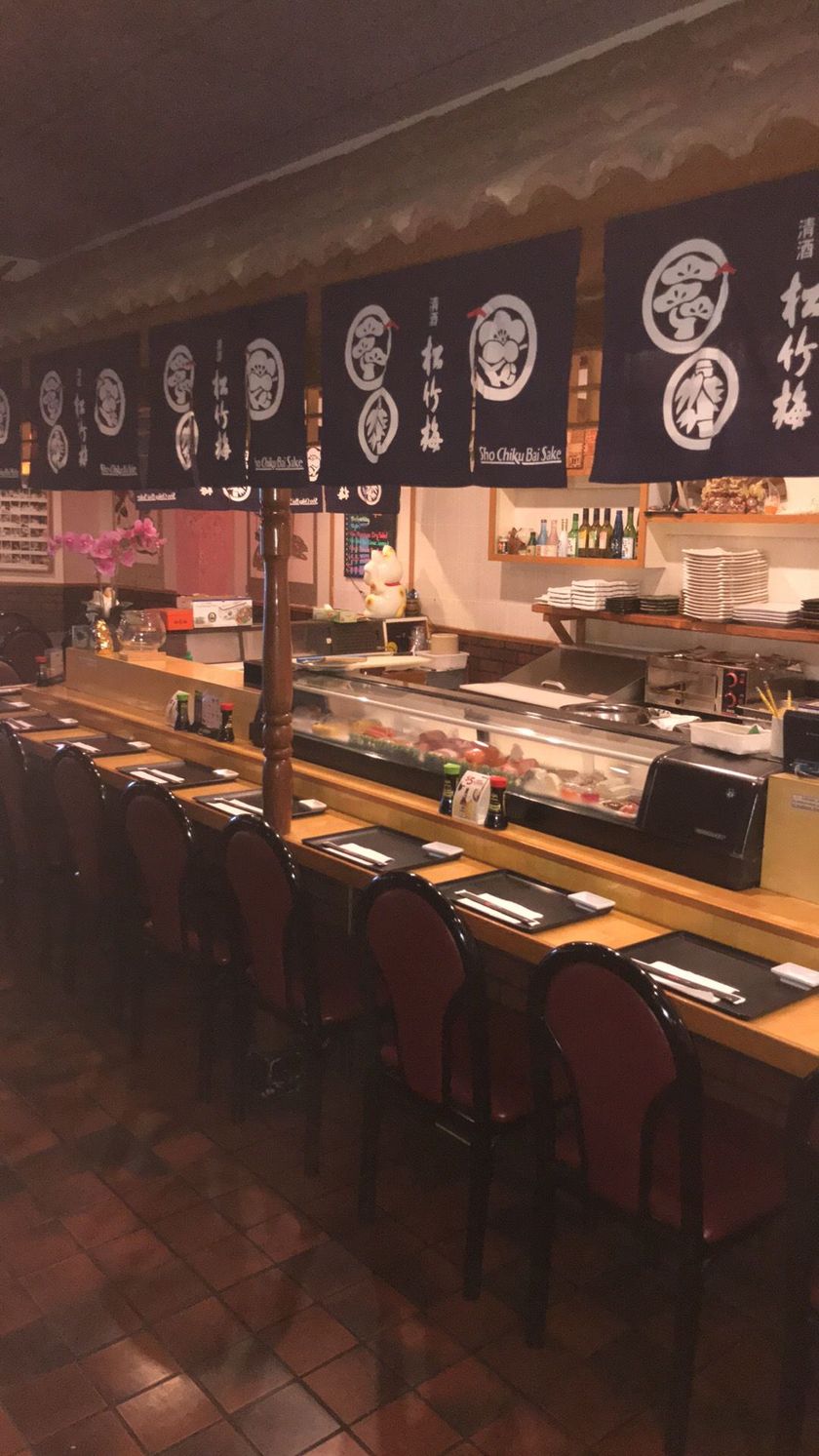 Empty sushi bar with counter seating, hanging black menu banners, and warm lighting.