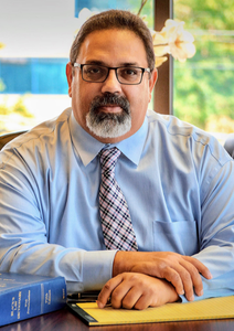 Man in glasses and blue shirt sitting at a desk with books, facing the camera, in a bright office setting