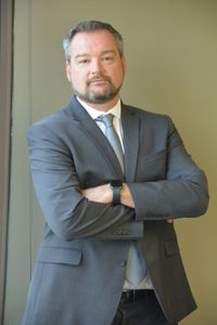 Man in a gray suit with arms crossed, standing against a plain green wall.