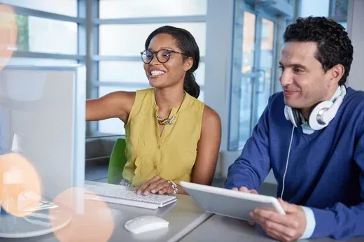 A man and a woman are sitting at a desk looking at a laptop computer.